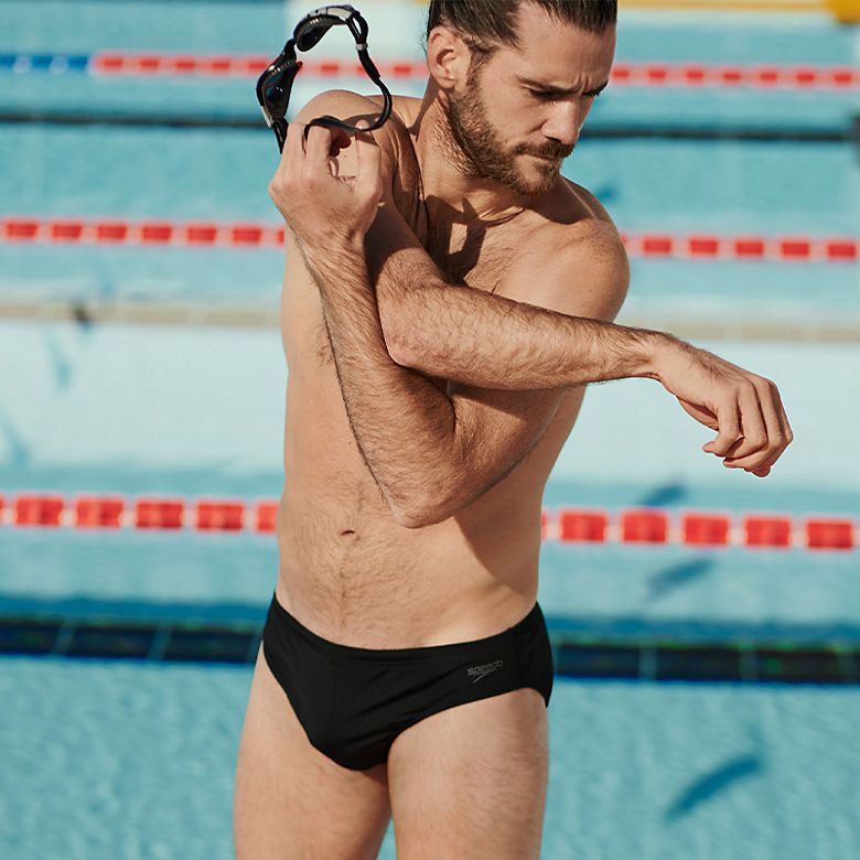 Man wearing black Speedo swim briefs, stretching beside indoor pool 