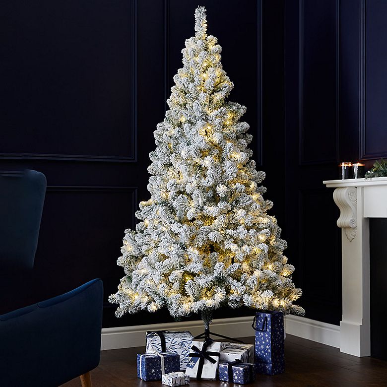 Snowy pre-lit Christmas tree with presents in a dark blue room with velvet chair. Shop Christmas trees 