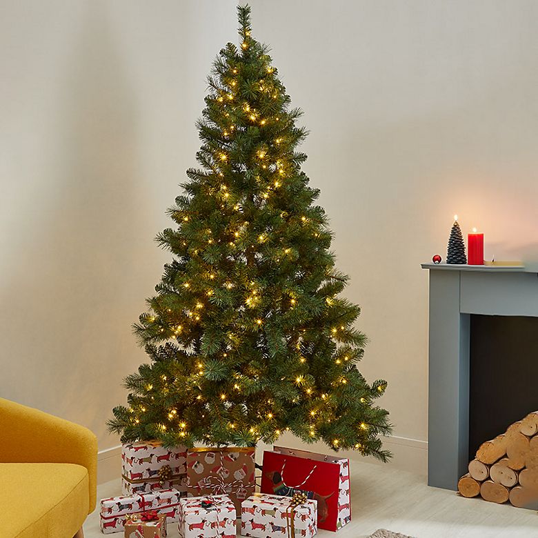 Lit Christmas tree with presents in room with mustard armchair. Shop Christmas trees