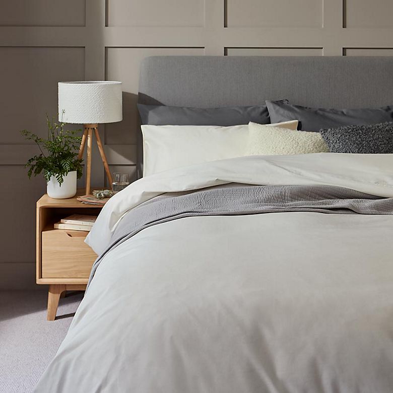 Grey Body Sensor bedding on a grey bed scattered with cream and grey cushions beside a wooden bedside table and lamp