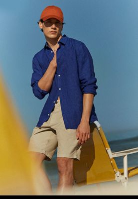 Man wearing a cobalt blue linen long-sleeved shirt, red cap and stone chino shorts. Shop the linen shirt