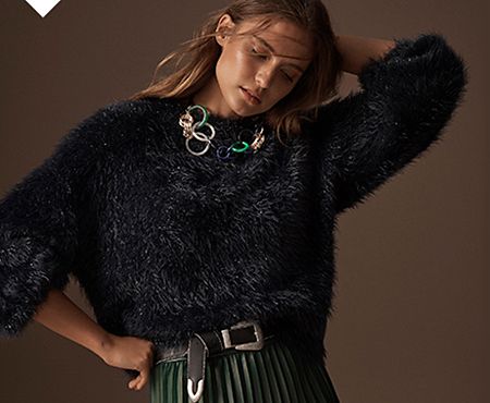 Model wears a navy faux-fur jumper over a green pleated faux-leather midi skirt with a black belt and statement necklace
