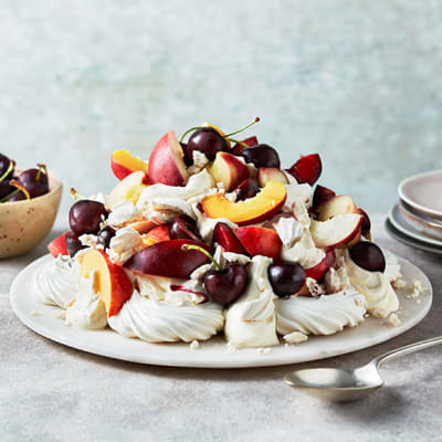A pavlova topped with stone fruits, with a bowl of cherries in the background