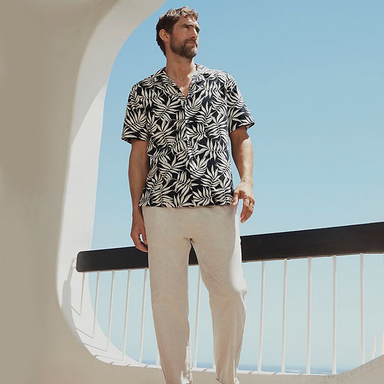 Man wearing summer holiday clothing including a printed shirt and linen trousers. Shop men’s holiday clothing