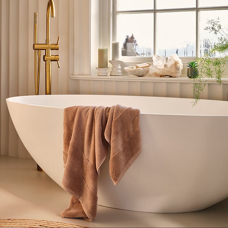 White bathtub with brass taps and a blush pink towel draped over the side. Shop the spa and wellness range
