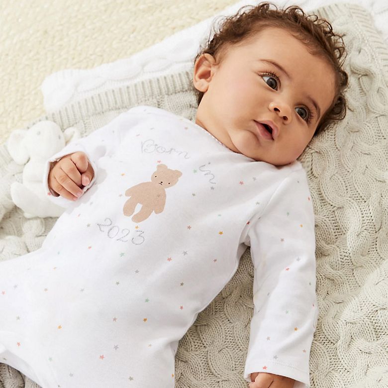 Baby wearing white sleepsuit with ‘born in 2023’ design. Shop babygrows.