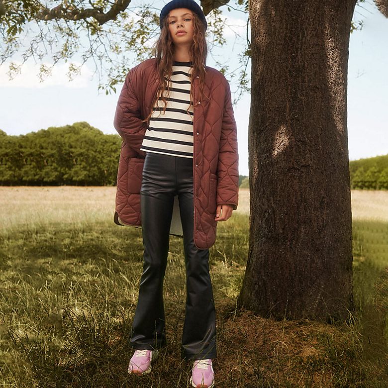 Girl wearing a long brown quilted jacket, striped top and black flares in a field. Shop kids’ coats 