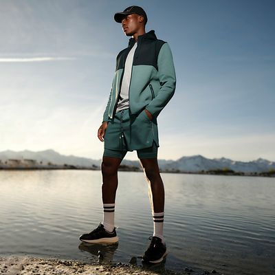 Man by a lake wearing workout shorts, a zip-up jacket, trainers and a baseball cap. Shop Goodmove activewear