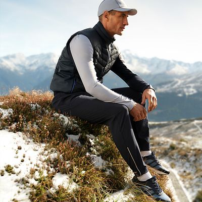 Man sitting on a snowy hill wearing workout top, leggings, padded gilet, trainers and baseball cap. Shop Goodmove activewear