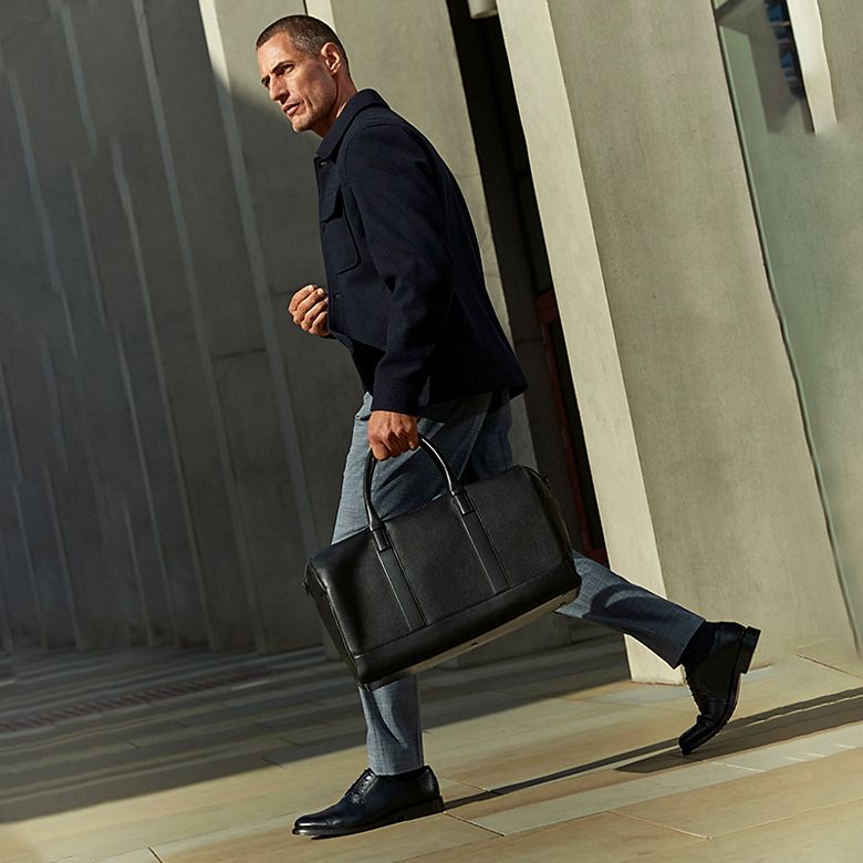 Man wearing a navy jacket and grey trousers, carrying a black leather overnight bag. Shop men’s smart wear 