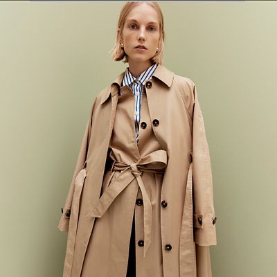 Woman wearing layered camel trench coats and a blue and white striped shirt. Shop women’s trench coats 
