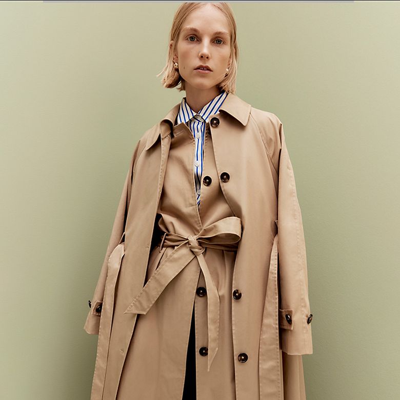 Woman wearing layered camel trench coats and a blue and white striped shirt. Shop women’s trench coats 