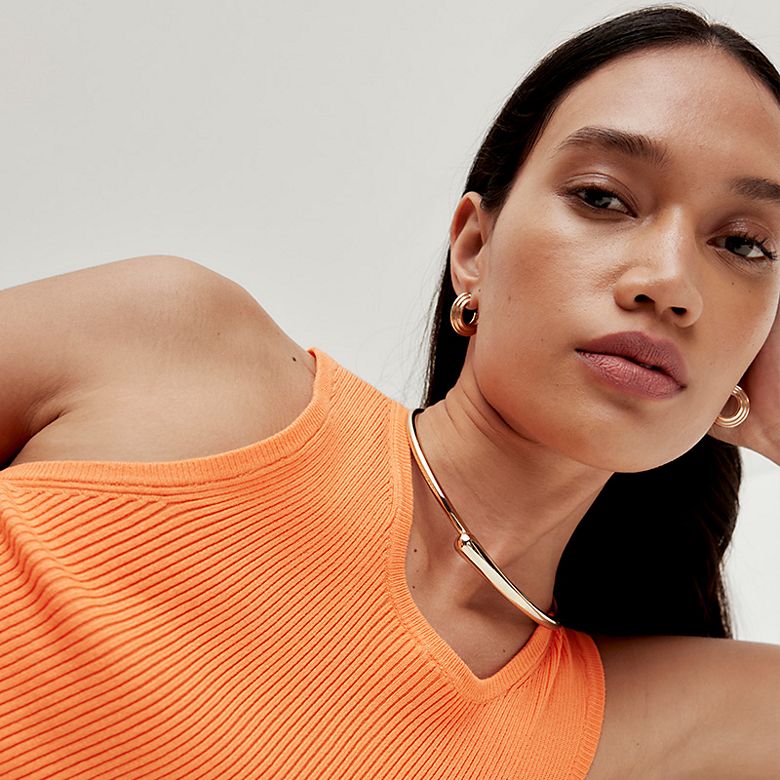 Woman wearing chunky hoop earrings and a choker necklace with a tangerine tank top. Shop jewellery 