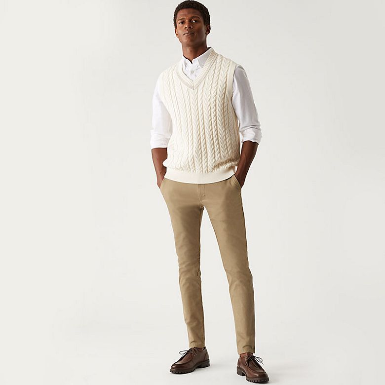 Man wearing beige skinny chinos, white shirt and cream cable-knit vest. Shop skinny chinos