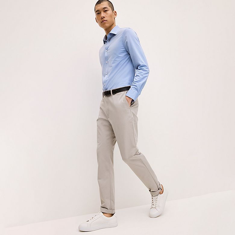 Man wearing grey slim-fit chinos, a blue shirt and white trainers. Shop slim-fit chinos