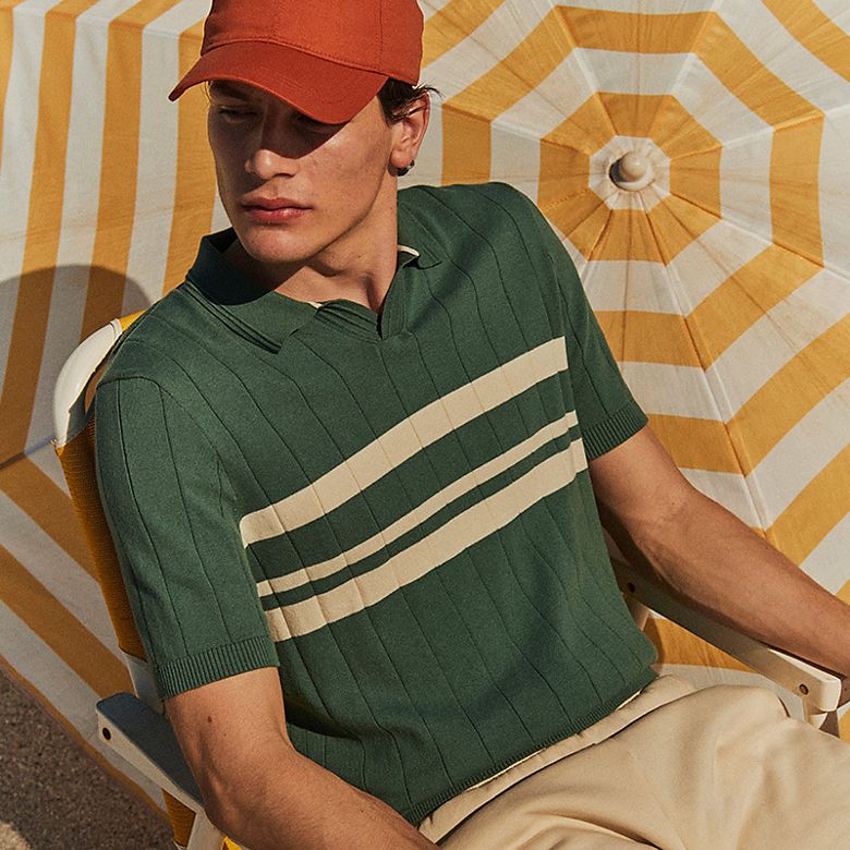 Man wearing an orange cap, green and white striped polo shirt, and ecru jersey shorts. Shop men’s shorts