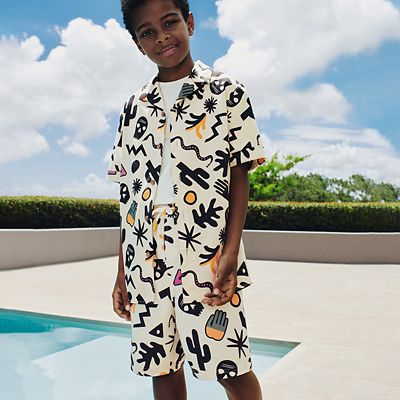Boy wearing a patterned shirt and matching shorts by a swimming pool. Shop boys’ summer clothing 