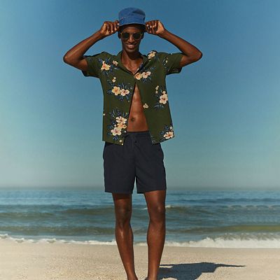 Man wearing summer holiday clothing including a printed shirt and navy swim shorts. Shop men’s holiday clothing