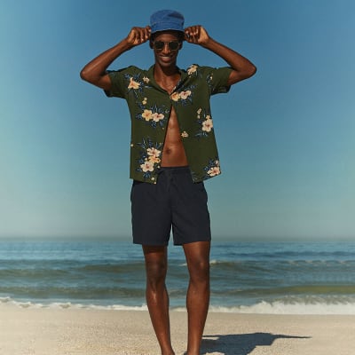 Man wearing summer holiday clothing including a printed shirt and navy swim shorts. Shop men’s holiday clothing