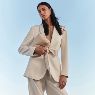 Woman wearing gold earrings, ivory belted jacket, beige top and wide-leg trousers. Shop modest womenswear