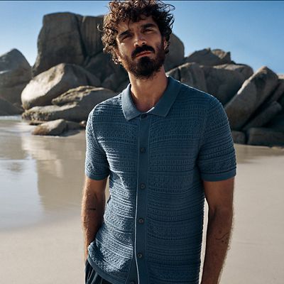 Man wearing blue short-sleeved collared cardigan at the beach. Shop men’s knitwear