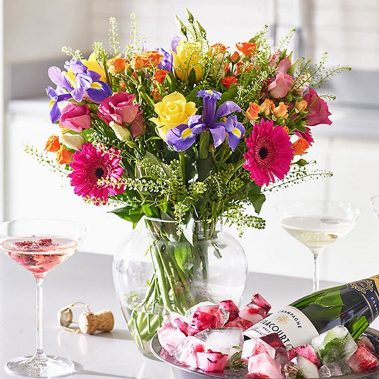 Vase of colourful flowers, including yellow and pink roses, irises and cerise germini, and a bottle of champagne