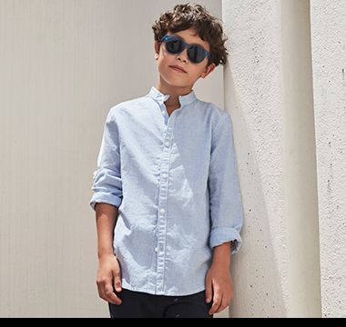 Boy wearing light blue shirt, navy chino shirts and sunglasses. Shop boys’ shirts