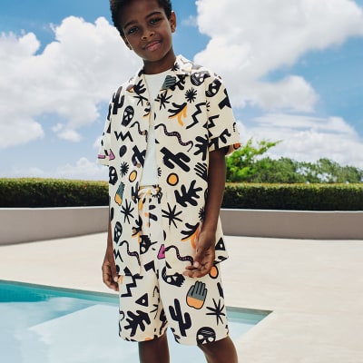 Boy wearing a patterned shirt and matching shorts by a swimming pool. Shop boys’ summer clothing 