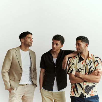 Man wearing a white top with beige blazer and chinos, another man wearing a white top under a black short-sleeved shirt and beige chinos and a third man wearing a multi-coloured graphic print short-sleeved shirt and blue jeans. Shop all men’s brands
