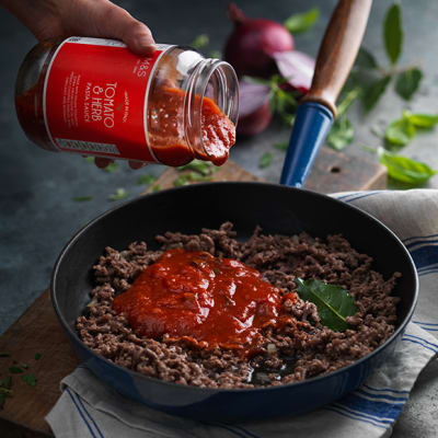 A jar of tomato and herb sauce being poured over sauteed beef mince