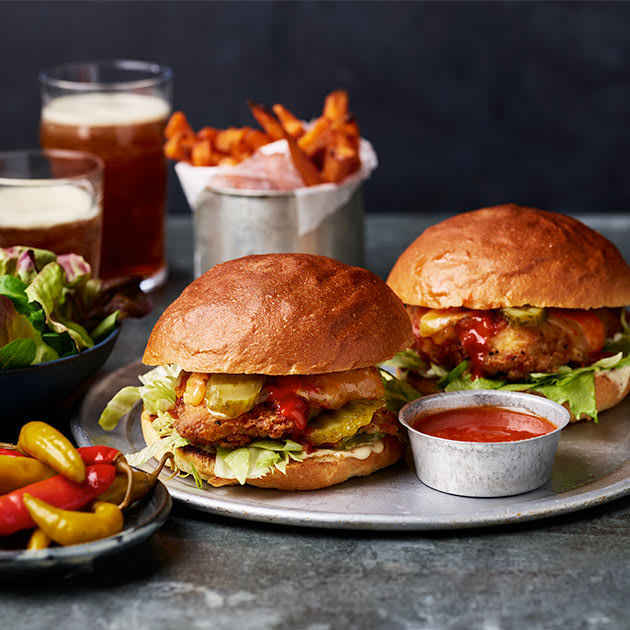 Two buffalo chicken burgers on a plate with a pot of buffalo sauce for dipping and some sweet potato fries in the background