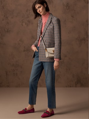 Model wears a black and white checked blazer over a pink cable-knit sweater and blue jeans with a white cross-body bag  and red loafers