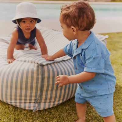 Baby wearing a navy romper and blue and white-striped bucket hat and a toddler wearing a light blue short-sleeved shirt and matching shorts. Shop new in baby