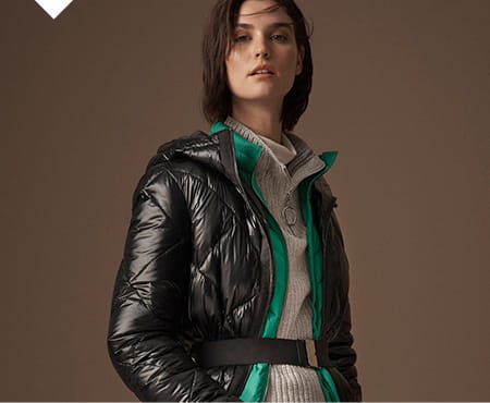 Model wears a black padded coat over a green padded jacket and grey ribbed jumper